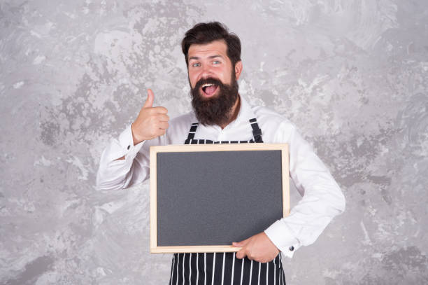 istock cook show empty board. thumb up for success. best chef menu. chalkboard with copy space for text. professional baker in apron. happy barista with beard. advertisement and food 1366967446
