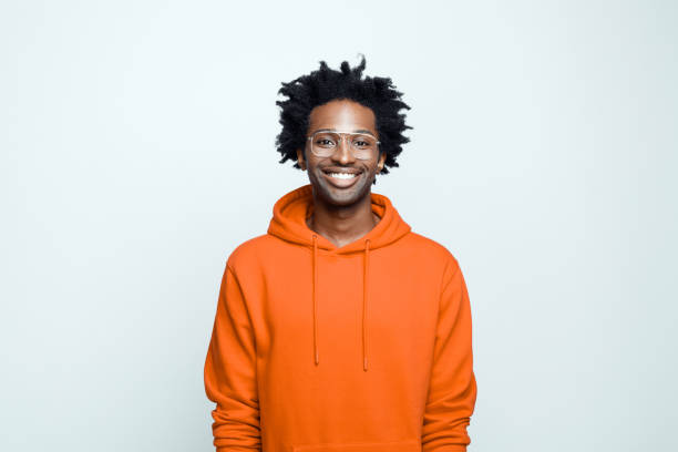 Portrait of cheerful man in orange hoodie Happy afro american young man wearing orange hoodie and glasses, smiling at camera. Studio shot on grey background. men hairstyle afro cool stock pictures, royalty-free photos & images