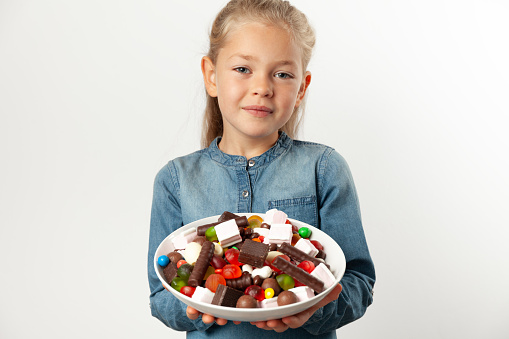 An 8-year-old Caucasian girl holds vegetables, red peppers and salad in her hands and thinks about what to choose, portrait on a light background.