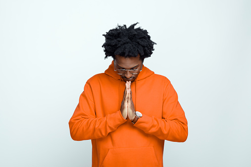 Afro american young man wearing orange hoodie and glasses, raising hand, closing eyes and meditating. Studio shot on grey background.