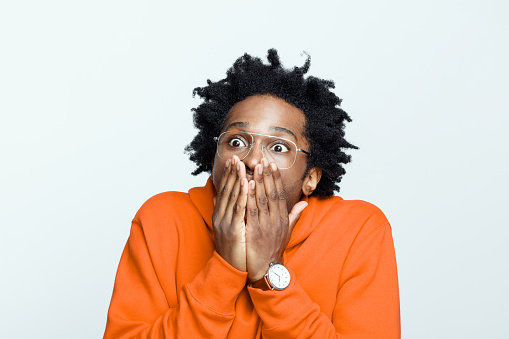 Shocked afro american young man wearing orange hoodie and glasses, covering mouth with hands. Studio shot on grey background.