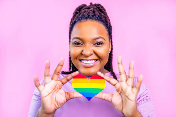 ritratto di un'attraente donna ispanica che tiene in mano un cuore arcobaleno lgbt in uno studio rosa - asexual reproduction foto e immagini stock