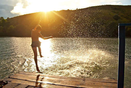 Children jump into the water from the pier. Camping by the water. Active rest on a sunset background. Concept of summer, vacation, travel and vacation. Water splashes on a background of sunlight.