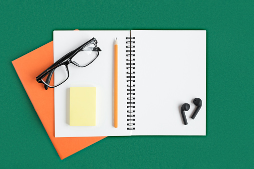 Notebook with pencil, glasses, wireless headphones. Open notebook on dark green color background, spiral notepad. Top view of notepad with blank page. Office flat lay. White Page for write. Copy Space
