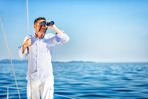 Handsome man with binoculars observing the sea on sailboat.