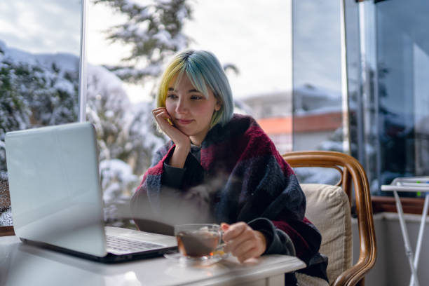 Young girl sitting on the balcony of her house while it is snowing and using laptop while enjoying the view and drinking tea stock photo