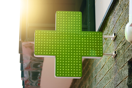 Pharmacy green light cross board. Drugstore signboard on building. Store decoration of downtown. Logo of city dispensary. Sunlight on background.