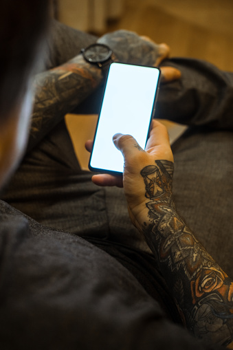 Man with tattoos using smart phone at home. Over the shoulder shoot. White screen visible and in focus.