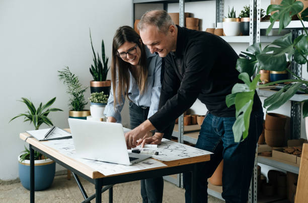 Happy Team of Architects Working Together on a New Business Projects Female and male colleague engineers leaning on the table and discussing a project design using blueprints and a laptop computer in their office. architect office stock pictures, royalty-free photos & images