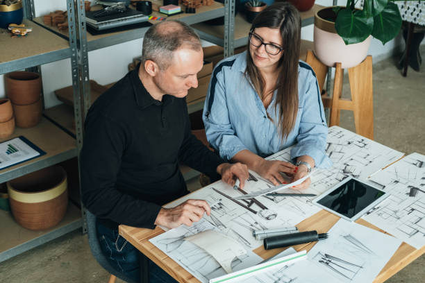 Happy Team of Furniture Designer Working Together on a New Business Projects Meeting Female and male colleague sitting at table and discussing a project design using blueprints and a digital tablet in their office. industrial designer stock pictures, royalty-free photos & images