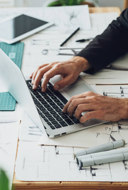 Close Up Photo of Architect Hands Using Laptop Computer in his Office High angle view of an anonymous furniture designer sitting at his desk and working with blueprints, sketches, digital tablet and laptop computer on a new business project. industrial designer stock pictures, royalty-free photos & images