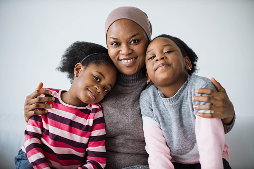Portrait of caring islamic african mother in hijab hugging her two cute daughters whiel sitting together on white cozy sofa. Concept of family, love and lifestyles.