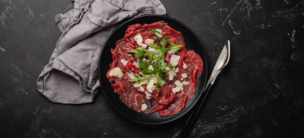 Cold meat appetizer Beef carpaccio with parmesan cheese and arugula on black plate Italian cold meat appetizer Beef carpaccio with parmesan cheese and arugula on black plate with fork and knife on dark stone concrete rustic background flat lay from above carpaccio parmesan cheese beef raw stock pictures, royalty-free photos & images