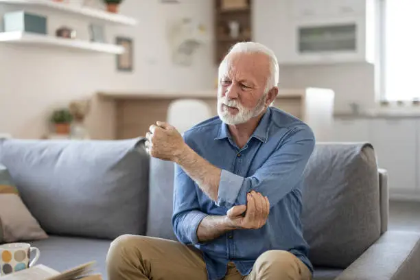 Senior man with arm pain.Old male massaging painful hand indoors. Old man hand holding his elbow suffering from elbow pain. Senior man suffering from pain in hand at home. Old age, health