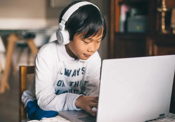 Asian boy with laptop and headphones, study from home. He is happy because he communicates with his friends. On his sweater it is written "always in the clouds"