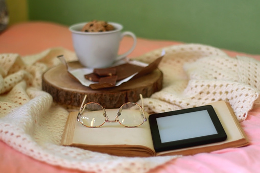 Mug of chocolate chip cookies, bar of chocolate, open book, reading glasses and e-reader on a bed. Selective focus.