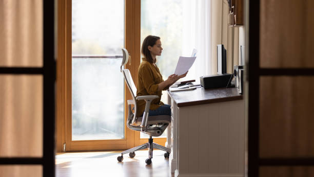 Serious home distance employee doing paperwork at table Serious home distance employee doing paperwork at table, reviewing financial paper reports at desktop computer, working at comfortable safe workplace with ergonomic office chair. Full length shot ergonomics photos stock pictures, royalty-free photos & images