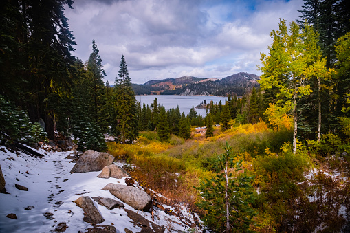 A high elevation, Backcountry, lake near Lake Tahoe called Marlette Lake is surrounded by vibrant fall colors and a light dusting of fresh snow. To the left of the image a snow covered trail with a single set of footprints can be seen.