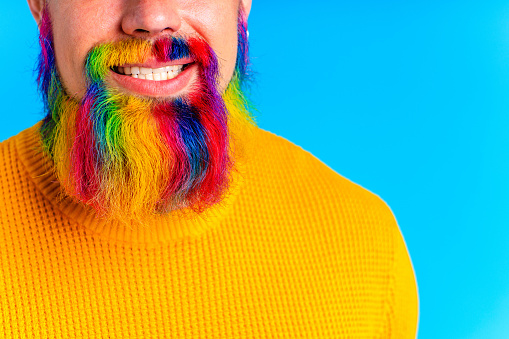 handsome man with raindow in beard and yellow sweater in blue studio baclground.