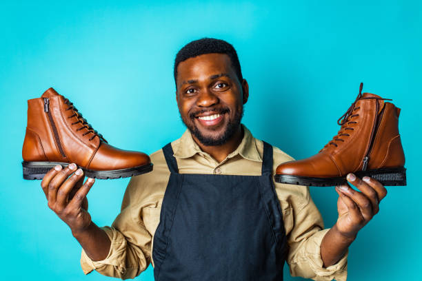 latin hispanic man in black apron showing brown leather shoes in blue studio background latin hispanic man in black apron showing brown leather shoes in blue studio background. cobbler stock pictures, royalty-free photos & images