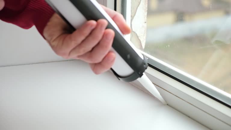 A man applies a sealant to the seam between the window and the sill from a tube of silicone sealant caulking with gun.