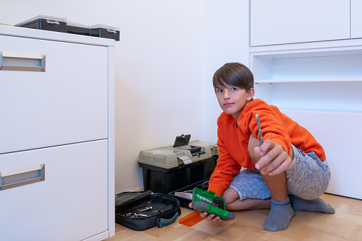 Satisfied brunette teenager in a orange sweater assembles white cabinets in his youth new room and helps himself with tools (hammer, screwdriver, drill bit). The boy looks at the camera.