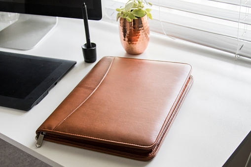 Brown leather business padfolio portfolio folder on sunny white office desk with computer and tablet