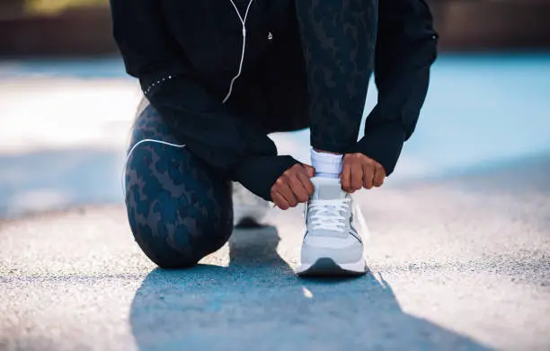 Cropped photo of a anonymous female athlete crouching to adjust her shoe for running. She is wearing black tights and white running shoes.