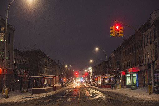 The entire roadway is covered with snow. The snow is not cleaned. An intersection is visible in the foreground and both, green and red traffic lights are on. The light from the traffic light illuminates the falling snow in red and green. Traces of cars are visible on snow. Cars parked at the sides of the street are also covered with snow. Snow continues to fall and snowflakes are visible flying in the air. Winter storm in progress and snow gathers in drifts. Early Morning. February 01, 2021. NYC. Brooklyn. Bay Ridge. New York. USA