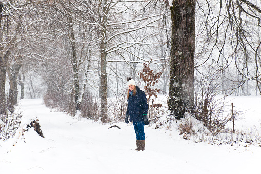 Woman with knit hat and long blond hair standing at a walking path in natural park at heavy snowfall. This image is part of a series.