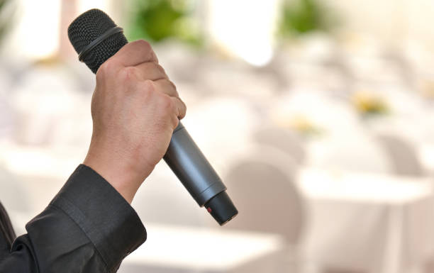 gros plan d’une main masculine tenant un microphone dans une salle de banquet ou une salle de bal décorée, vue latérale avec mise au point sélective et arrière-plan flou - spot lit photos et images de collection