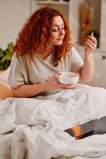 ベッドで朝食を食べる赤毛の女の子。寝室のベッドに座って、ラップトップでインターネット上でサーフィンしながらシリアルを食べる生姜の女の子。 - comfort food ストックフォトと画像