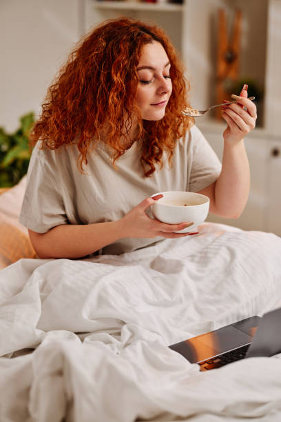 침대에서 아침 식사를 먹는 빨간 머리 소녀. 침실에 있는 침대에 앉아 노트북으로 인터넷에서 서핑을 하는 동안 시리얼을 먹는 생강 소녀. - comfort food 뉴스 사진 이미지