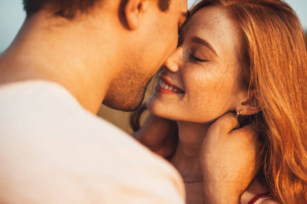 Close-up view of a man kissing his freckled girlfriend standing against a bright background of the afternoon sun. Love is in the air. Closeup portrait. Summer vacation. stock photo