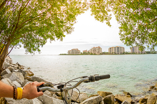 POV Point of view shot of a young latin sportsman barefoot biking a bicycle in front of a touristic Art Deco District landscape at South Pointe Park in Miami Beach, Miami, South Florida, United States of America - USA. On background, the famous, exclusive and luxury Fisher Island. Shooting from a personal perspective in an exotic tropical beach travel holidays.