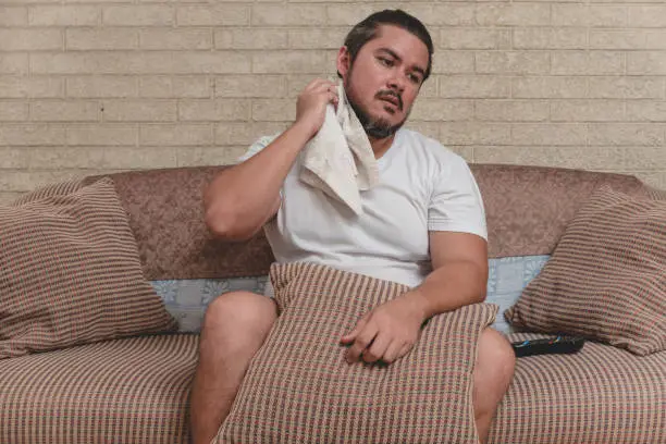 Photo of An overweight man wipes the sweat off his neck with a white towel. Heat wave, broken air conditioner or sweltering temperature indoors.