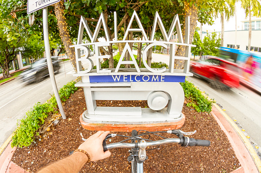 POV Point of view shot of a young latin sportsman biking a bicycle in front of a touristic Art Deco District landscape at Ocean Drive, South Beach, Miami Beach, Miami, South Florida, United States of America - USA. On background, the famous and touristic Art Deco District. Shooting from a personal perspective in an exotic tropical beach travel holidays.