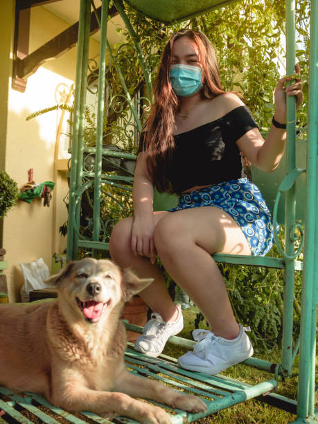 A cheerful young asian woman wearing a face mask sits on the glider swing with her faithful dog. New normal concept. A cheerful young asian woman wearing a face mask sits on the glider swing with her faithful dog. New normal concept. hot filipina women stock pictures, royalty-free photos & images
