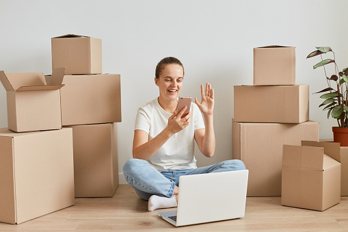 Image of positive woman mover wearing white T-shirt sitting on the floor near cardboard boxes, posing in front of laptop and having video call via cell phone, waving hand, saying hello.