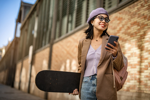 A young woman, a young Chinese woman, leads an urban life, carries her skateboard in her hands and uses a mobile phone. Hipster youth culture.