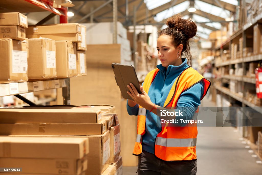 A warehouse worker takes inventory in the storage room. A warehouse worker is standing next to a shelf and using a digital tablet. Warehouse Stock Photo