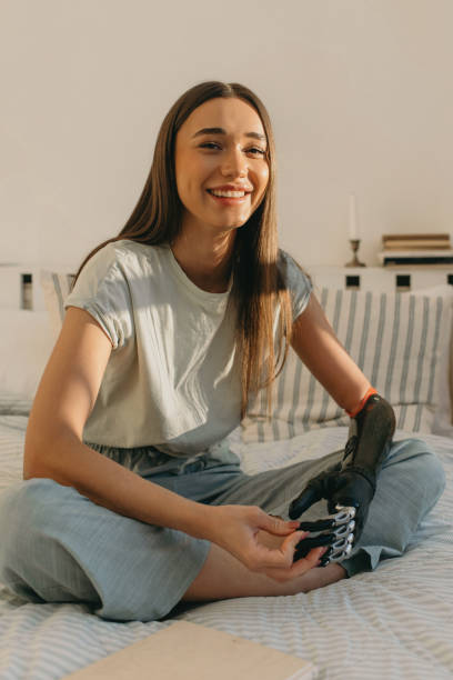 indoor portrait of beautiful laughing female with artificial black metal hand sitting on bed in casual pants and white t-shirt. technology and lifestyle. life after amputation. biomedical engineering - portrait vertical close up female imagens e fotografias de stock