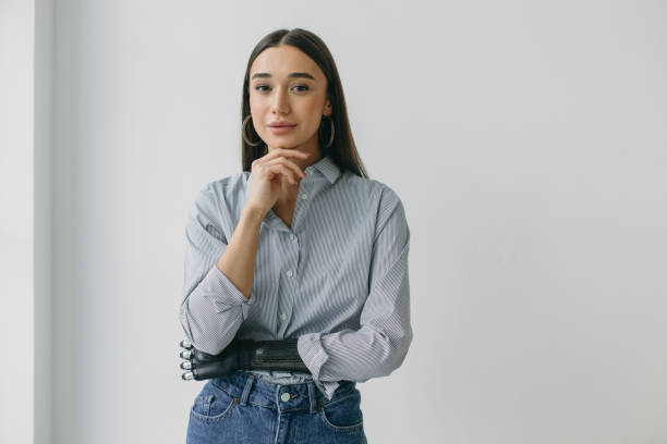 Horizontal portrait of shy gorgeous young lady in blue jeans and shirt holding chin, posing with bionic prosthetic arm, standing against white studio wall with copy space for your advertising content Horizontal portrait of shy gorgeous young lady in blue jeans and shirt holding chin, posing with bionic prosthetic arm, standing against white studio wall with copy space for your advertising content prosthetic equipment stock pictures, royalty-free photos & images