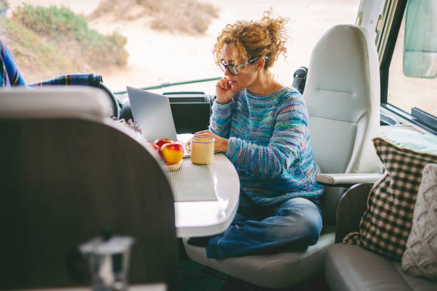 Adult nice woman work on laptop sitting in a camper van dinette enjoying freedom travel vacation or vanlife lifestyle. Modern job with computer connection technology. Female people freelance stock photo