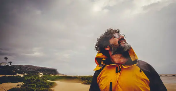 Close up of mature man enjoying bad weather and explore the outdoor beach. Cloudy and rainy sky. Happy free male people with yellow jacket smile and lok up