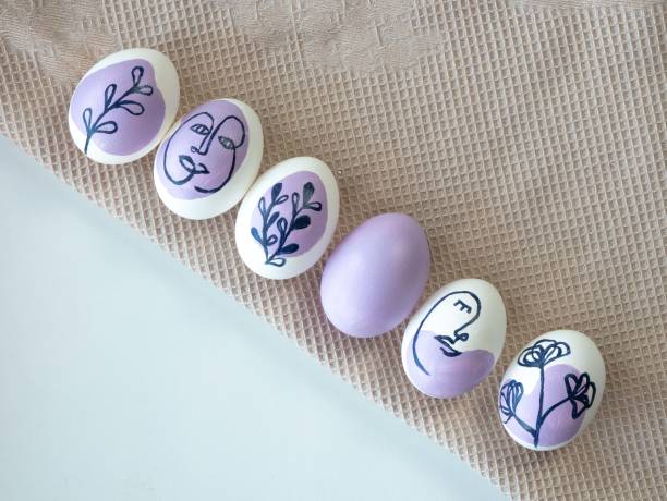 Beautiful Easter colored eggs on a kitchen towel lie in a row from corner to corner. The eggs are painted with a floral ornament and a human face in purple. Place for text.
