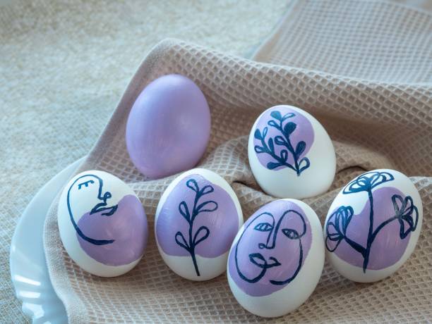istock Beautiful Easter colored eggs on a kitchen towel, on a white plate, next to green sprigs of grass and leaves. The eggs are painted with a floral ornament and a human face in purple 1367113816