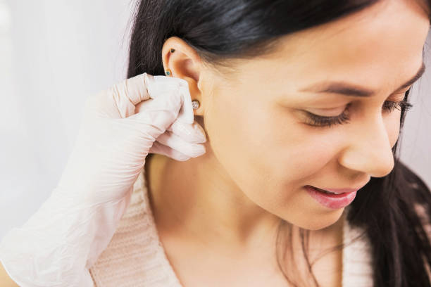 foto vista laterale di una bella donna bruna che si prepara a una procedura di piercing e medico in guanti sterili che strofina l'orecchio con l'alcol. - pierced foto e immagini stock
