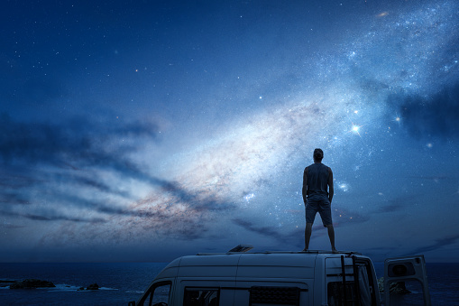 Night time with a beautiful starry sky. A man is standing on top of the roof of a camper van that's parked near the ocean.