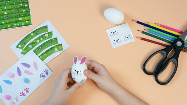 istock Children's hands make an Easter decoration on the egg. Pencils and stickers on a yellow background. Easter concept. 1367125780